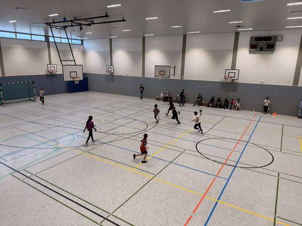 Das Bild zeigt eine Turnhalle von innen, in der viele Kinder ein Ballspiel spielen. Am Rand sitzen weitere Kinder und trinken.
