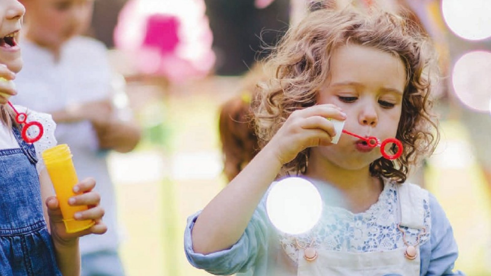 Ein fröhliches Mädchen steht im Vordergrund und bläst Seifenblasen, die in der Sonne schimmern und durch die Luft schweben. Im Hintergrund sind andere Kinder zu sehen, die lachen und spielen, was eine lebendige und fröhliche Atmosphäre schafft. 