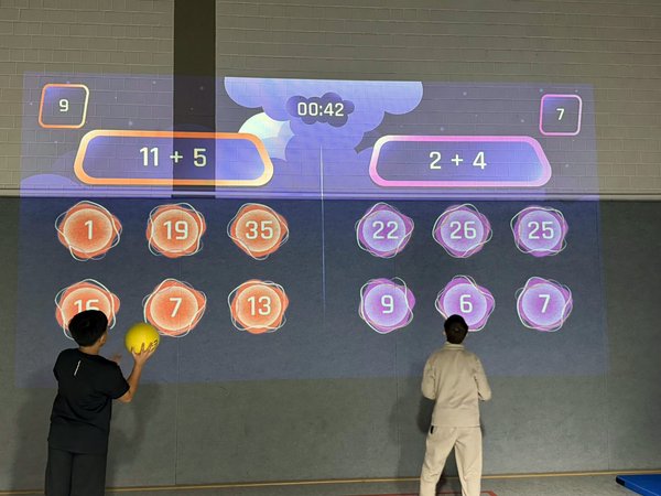 Das Bild zeigt zwei Kinder vor einer Wand auf der ein Rechenspiel projiziert ist. Mit einem Ball müssen sie auf die richtige Lösung werfen.