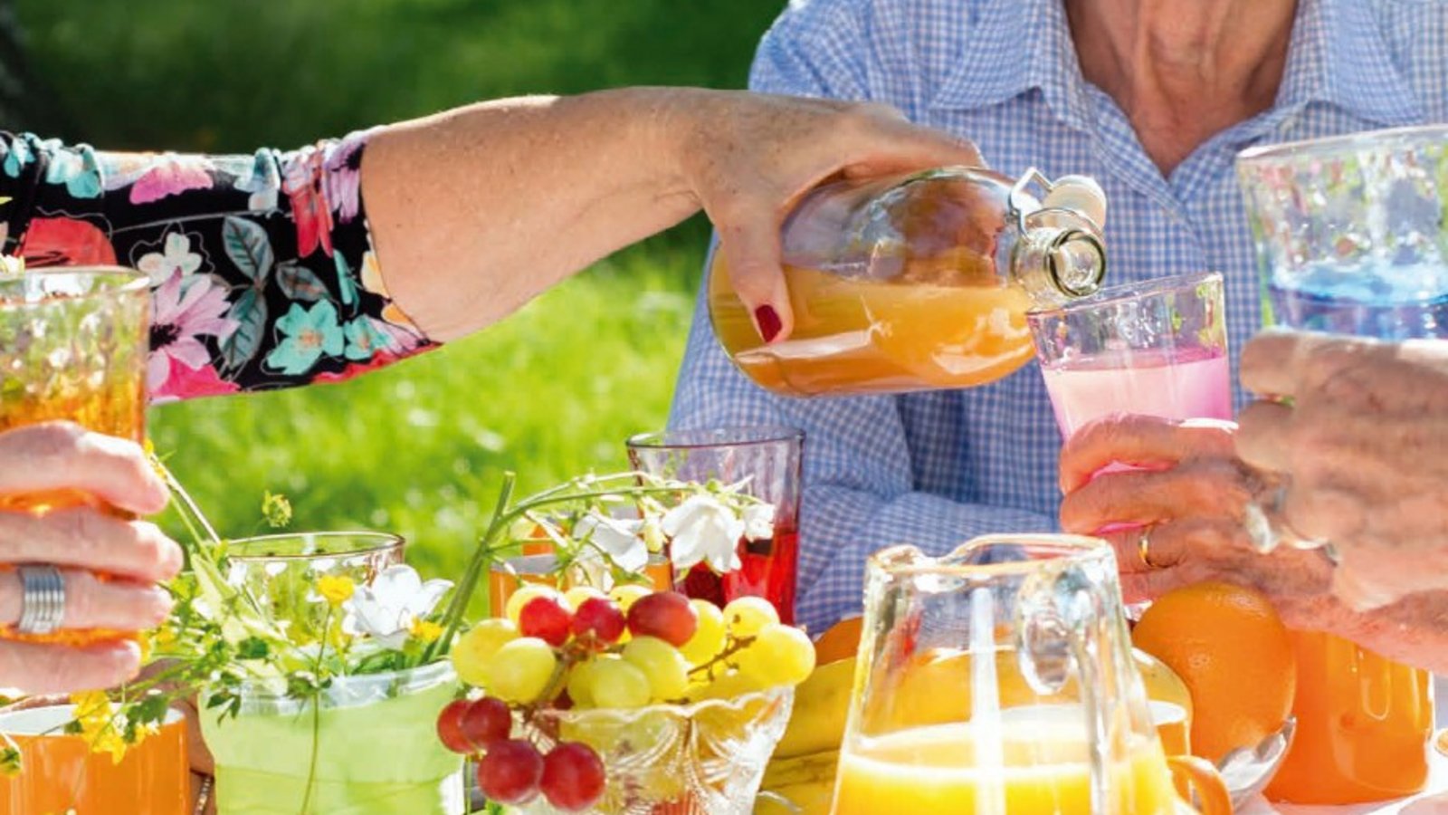 Ein gedeckter Tisch mit Trauben und Orangensaft. Eine Frau gießt einem Mann freundlich sein Glas ein.