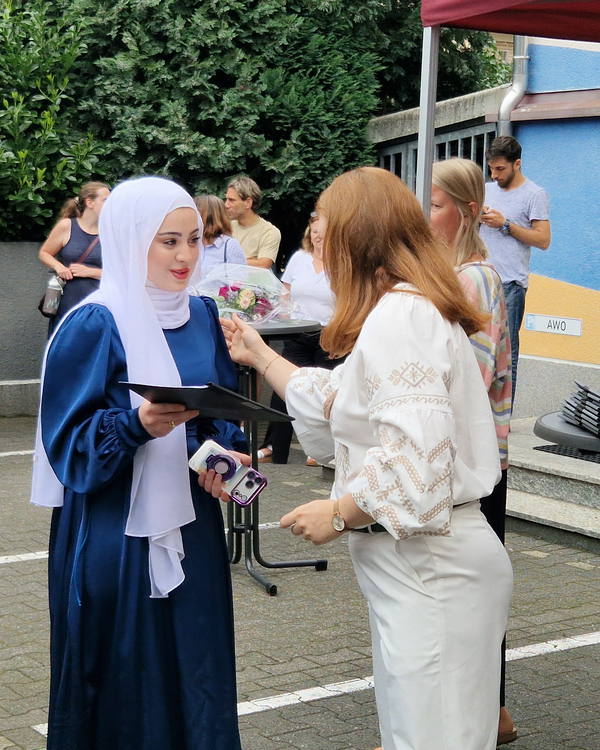 Auf dem Bild sieht man zwei Frauen, die miteinander sprechen. Links hat die Dame ein Kopftuch und hält ihr Zeugnis in der Hand. Die rechte Frau gratuliert ihr.